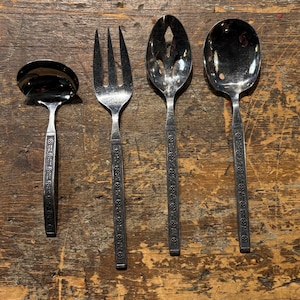 May include: A collection of four vintage silver serving utensils: a ladle, a fork, a slotted spoon, and a serving spoon. Each piece features a decorative handle pattern. The utensils are displayed on a rustic wooden surface.