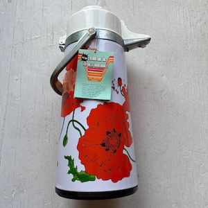 May include: A vintage thermos flask with a white body, decorated with large red poppy flower illustrations and green leaves. It has a white lid, a metal handle, and a black base. A tag reads "NEW THERMO JUG FOR FULLY UNDER-TABLE".