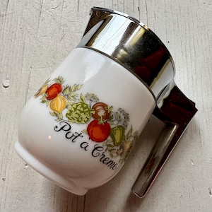 Puede incluir: Jarra blanca de cerámica "Pot a Creme" con asa y borde de metal plateado. La jarra está decorada con ilustraciones coloridas de verduras, como tomates, pimientos y alcachofas. El texto "Pot a Creme" está escrito en escritura negra.
