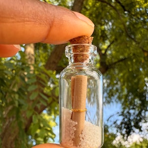May include: A small, clear glass bottle with a cork stopper, containing a rolled-up scroll, a gold ring, and white crystals. The bottle is held in a hand against a blurred green background of leaves and branches.