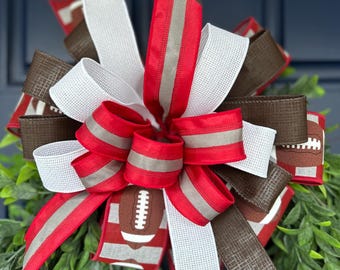 May include: A decorative bow featuring multiple ribbons in red, white, and dark brown. The ribbons include solid colors, a textured weave pattern, and a graphic print of brown footballs with white laces, arranged in a layered, full design.