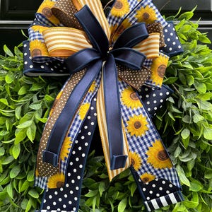 May include: A decorative bow featuring a variety of ribbons in navy blue, brown, yellow, and white. The bow includes ribbons with sunflowers, polka dots, stripes, and a gingham pattern. The bow is attached to a green wreath.