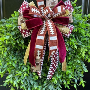 May include: A decorative wreath featuring lush green foliage adorned with a large, multi-layered bow. The bow consists of ribbons with football patterns, solid maroon velvet, white polka dots on maroon, and shimmering gold glitter accents.