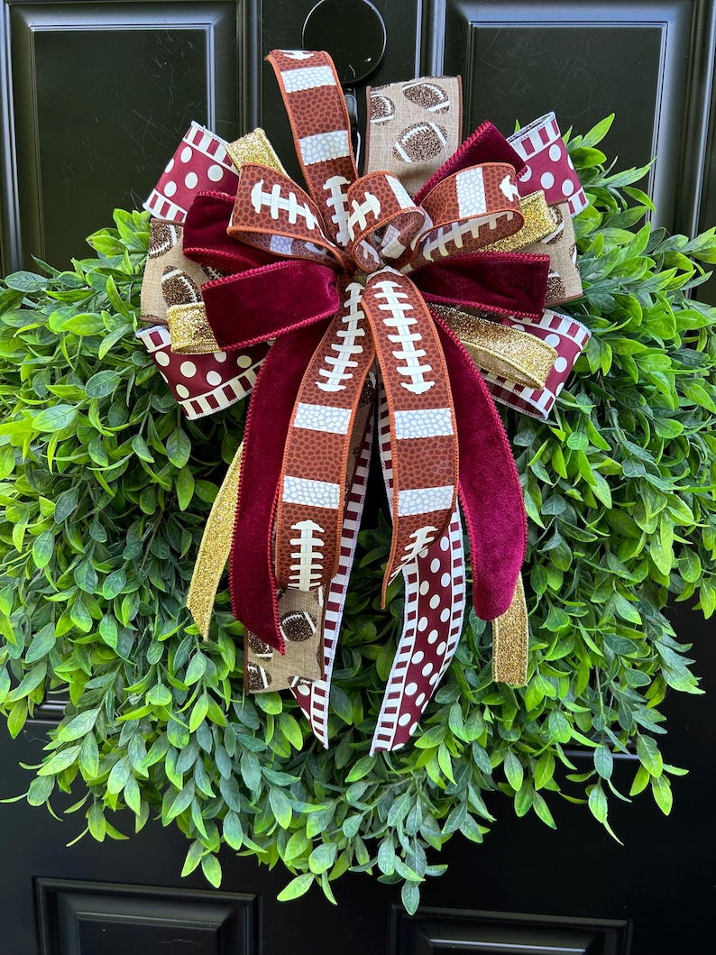 May include: A decorative wreath featuring lush green foliage adorned with a large, multi-layered bow. The bow consists of ribbons with football patterns, solid maroon velvet, white polka dots on maroon, and shimmering gold glitter accents.