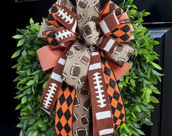 May include: A decorative bow featuring a mix of ribbons with football patterns, black and orange diamond prints, and solid orange textures, layered over a lush green leaf wreath. The ribbons display white football laces.