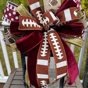 May include: A decorative bow featuring a mix of ribbons with football-themed patterns, solid deep red velvet, and gold glitter. The ribbons display brown football textures, white laces, and polka dots, arranged in a multi-layered, full design.