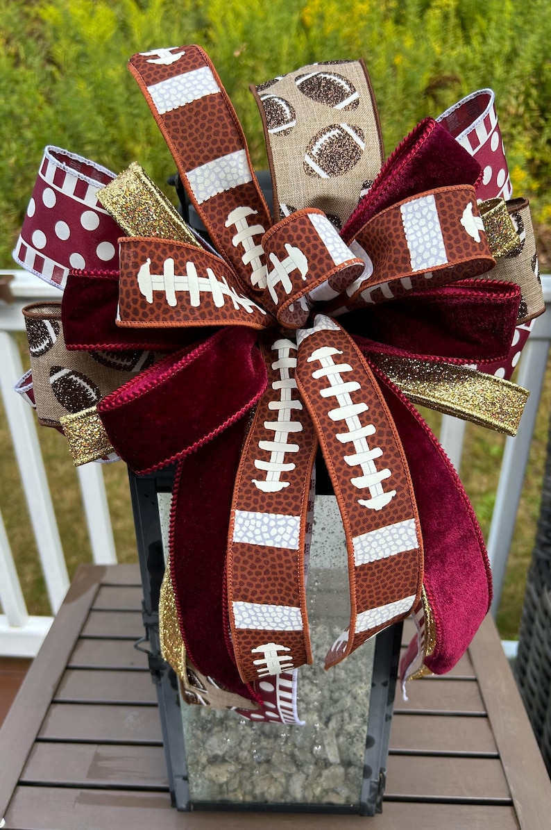 May include: A decorative bow featuring a mix of ribbons with football-themed patterns, solid deep red velvet, and gold glitter. The ribbons display brown football textures, white laces, and polka dots, arranged in a multi-layered, full design.
