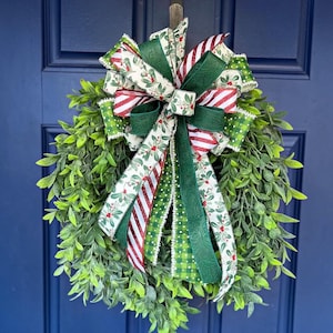 May include: A round green boxwood wreath featuring a large decorative bow made of multiple ribbons. The ribbons include patterns of red and white diagonal stripes, green and white polka dots, and a white background with holly berries.
