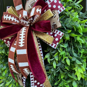 May include: A decorative bow featuring ribbons with football patterns, maroon velvet, and gold glitter, attached to a lush green leaf wreath. The ribbons display brown footballs with white laces and white polka dots on a maroon background.