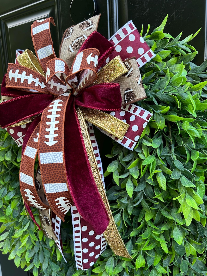 May include: A decorative bow featuring ribbons with football patterns, maroon velvet, and gold glitter, attached to a lush green leaf wreath. The ribbons display brown footballs with white laces and white polka dots on a maroon background.