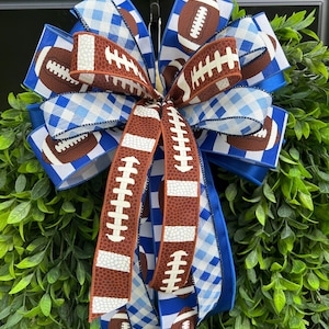 Puede incluir: Un lazo decorativo que combina cintas con gráficos de balones de fútbol americano en marrón, patrones de cuadros vichy en azul y blanco, y rayas azul sólido. El lazo está formado por múltiples bucles de cinta con alambre y extremos largos.