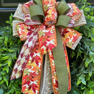 May include: A decorative bow featuring a mix of wired ribbons. The design includes a vibrant autumn leaf print in red, orange, and yellow, solid olive green fabric, a tan ribbon with white scroll patterns, and a burgundy and white diamond pattern.