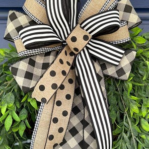 May include: A decorative wreath bow with various patterned ribbons. The bow incorporates black and white striped, polka dot, and buffalo check ribbons. The bow is set against a backdrop of green foliage.