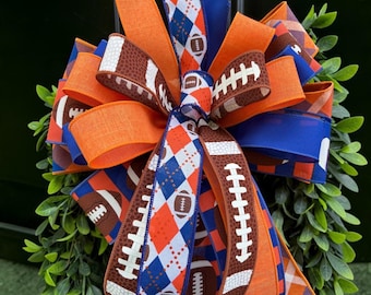 May include: A decorative bow made of multiple ribbons featuring orange, blue, and brown colors. The ribbons display patterns of American footballs, argyle diamonds, and solid orange or blue fabric, arranged in a multi-layered, full design.