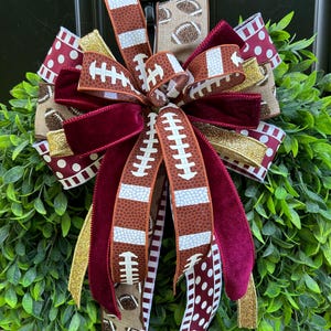 May include: A decorative bow featuring multiple ribbons with football patterns, maroon velvet, gold glitter, and white polka dots on a maroon background. The bow is layered with various textures and colors, creating a full, multi-loop arrangement.