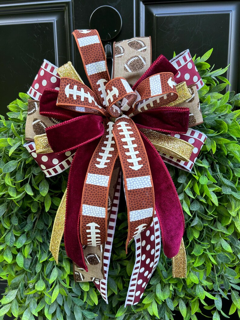 May include: A decorative bow featuring multiple ribbons with football patterns, maroon velvet, gold glitter, and white polka dots on a maroon background. The bow is layered with various textures and colors, creating a full, multi-loop arrangement.