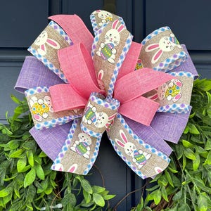 Pode incluir: Uma grinalda decorativa de Páscoa com um grande laço de fitas rosa e roxas. O laço apresenta uma fita estampada com coelhinhos e ovos de Páscoa. A grinalda é rodeada por folhas verdes, criando uma decoração primaveril festiva.