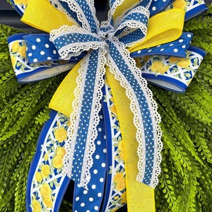 May include: A decorative bow featuring blue and yellow ribbons. The bow has a white lace trim and is adorned with white polka dots and yellow rose patterns. The bow is set against a backdrop of green foliage.