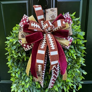 May include: A decorative wreath featuring a large, multi-layered bow made of ribbons with various patterns, including football motifs, maroon velvet, gold glitter, and white polka dots on a maroon background, centered on a lush green leaf base.