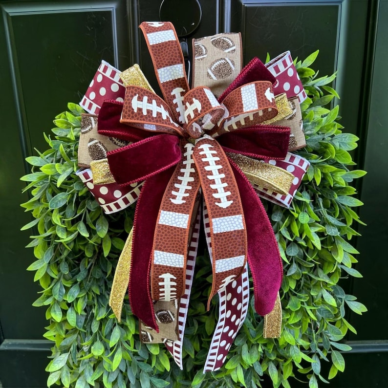 May include: A decorative wreath featuring a large, multi-layered bow made of ribbons with various patterns, including football motifs, maroon velvet, gold glitter, and white polka dots on a maroon background, centered on a lush green leaf base.