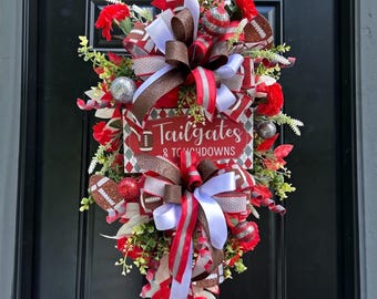 May include: A decorative door wreath featuring red, white, and brown ribbons with football patterns, red flowers, and silver glitter ornaments. A central rectangular sign displays the text Tailgates & Touchdowns in white script.