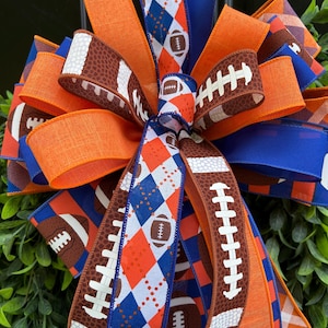 May include: A decorative bow made of wired ribbon featuring a mix of solid orange, solid blue, and patterned fabrics. The patterns include a blue and orange argyle print with football icons and a brown football texture with white laces.