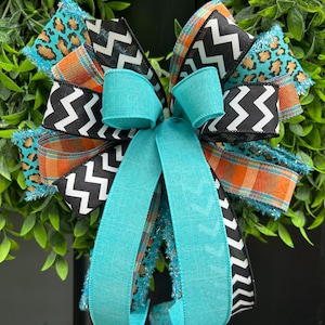 May include: A decorative bow featuring multiple ribbons with varied patterns including a turquoise leopard print, an orange and blue plaid, and a black and white chevron design. The center is accented with solid turquoise fabric ribbons.