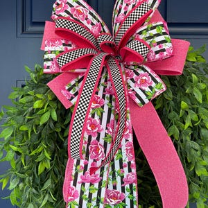 May include: A decorative bow for a wreath, featuring a striking combination of patterns and colours. The bow incorporates pink, black and white checkered, and striped ribbons, decorated with pink floral patterns. The bow is set against a backdrop of green foliage.