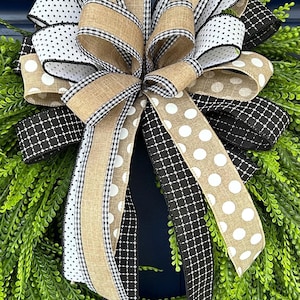 May include: A decorative bow featuring multiple ribbons with varied patterns including tan burlap, black and white polka dots, black and white gingham, and black fabric with a white cross-hatch design, all layered over a green artificial leaf wreath.