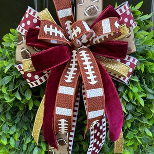 May include: A decorative bow made of multiple ribbons featuring football patterns, maroon velvet, gold glitter, and white polka dots on a maroon background. The ribbons are layered and tied together, creating a full, multi-textured arrangement.