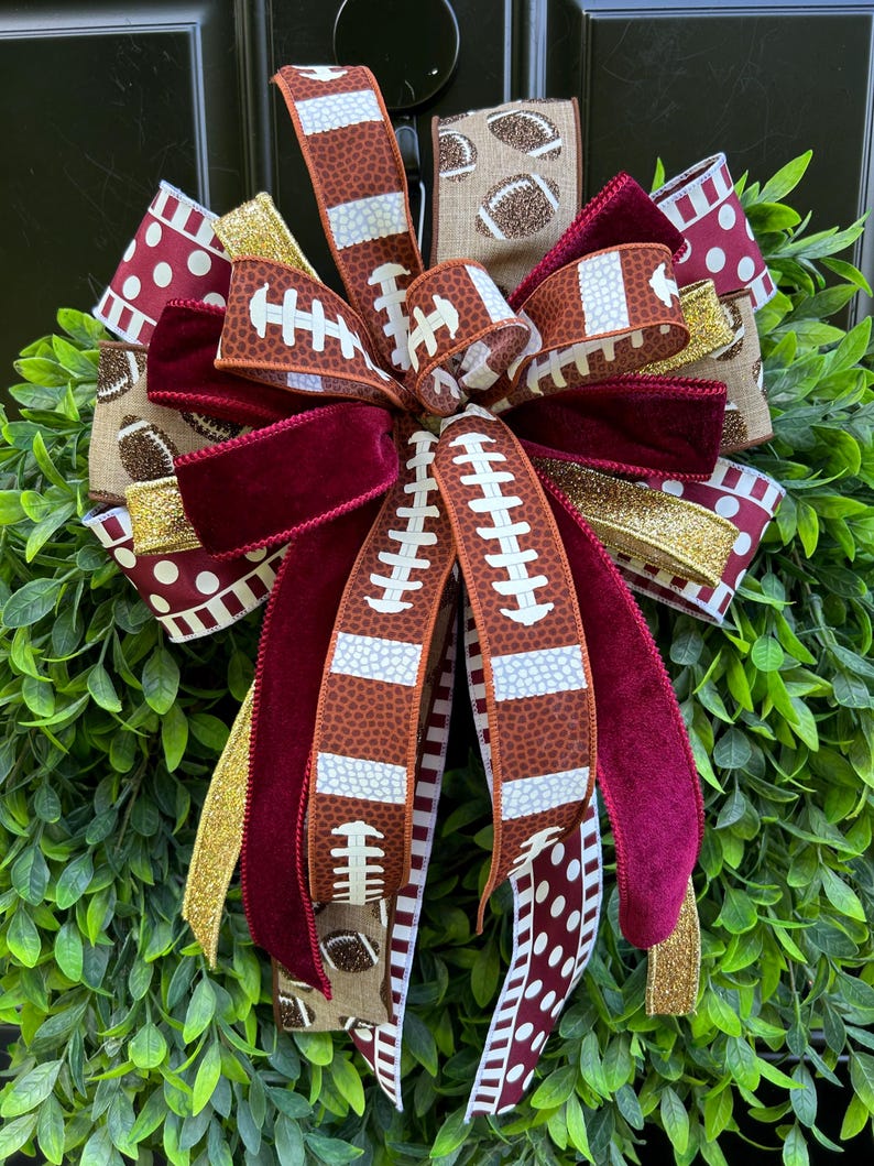 May include: A decorative bow made of multiple ribbons featuring football patterns, maroon velvet, gold glitter, and white polka dots on a maroon background. The ribbons are layered and tied together, creating a full, multi-textured arrangement.