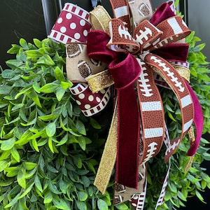 May include: A decorative wreath featuring lush green leaves adorned with a multi-layered bow. The bow includes ribbons with football patterns, maroon polka dots, solid maroon velvet, and shimmering gold glitter fabric accents.