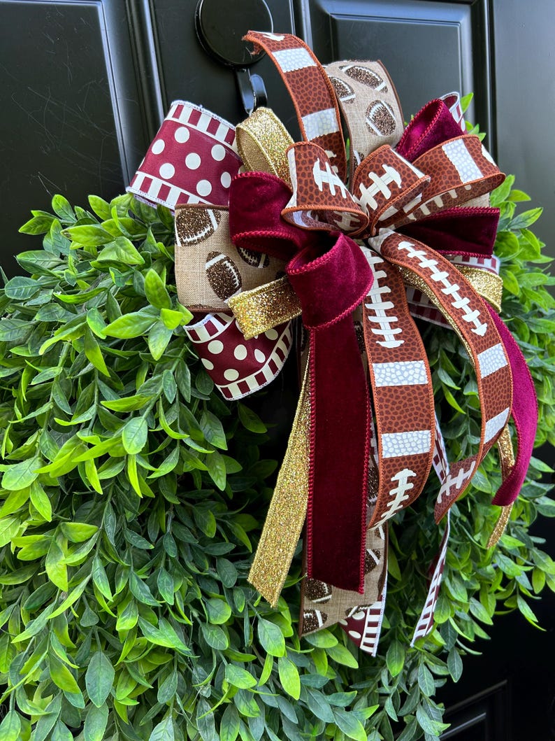 May include: A decorative wreath featuring lush green leaves adorned with a multi-layered bow. The bow includes ribbons with football patterns, maroon polka dots, solid maroon velvet, and shimmering gold glitter fabric accents.