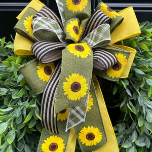 May include: A decorative bow with a sunflower design. The bow features layers of yellow, olive green, and black and white striped ribbons. The bow is attached to a green leafy base, creating a bright and cheerful aesthetic.