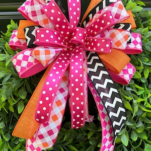 May include: A decorative bow made of multiple ribbons featuring a vibrant mix of patterns and colours. The ribbons include hot pink with white polka dots, orange solid fabric, black and white chevron stripes, and a pink, orange, and white plaid design.