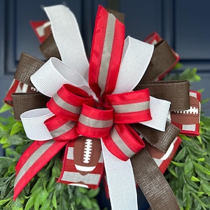 May include: A decorative bow featuring red, white, and brown ribbons. The red ribbons have a silver stripe, and some ribbons display a football design. The bow is set against a backdrop of green foliage.