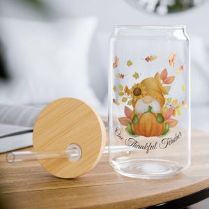Puede incluir: Un vaso transparente en forma de lata con tapa y pajita de madera. El vaso presenta un diseño de gnomo con temática otoñal con una calabaza y el texto "One Thankful Teacher". El diseño incluye hojas de otoño y un girasol.