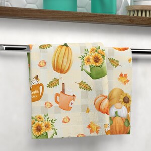 Puede incluir: Una toalla plegada con temática otoñal. Presenta un patrón repetido de calabazas, girasoles y hojas de otoño sobre un fondo a cuadros color crema. Un gnomo también está presente en el diseño. La toalla está colgada de una barra cromada.