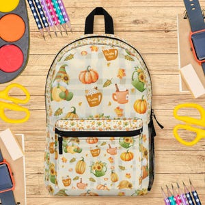 Puede incluir: Una mochila color crema con cremallera y asa negras, con un estampado de temática otoñal de calabazas, girasoles y tazas de café. La mochila tiene un bolsillo frontal y un bolsillo lateral de malla. El diseño incluye un gnomo y el texto "Pumpkin Spice".