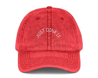 Puede incluir: Gorra de béisbol roja de estilo vintage con visera curva. La gorra presenta el texto bordado "JUST DINK IT" en blanco, arqueado en la parte delantera. El sombrero está hecho de un material suave y texturizado.
