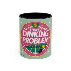 May include: A ceramic coffee mug with a black interior and a light green exterior. The mug features a pink and white graphic with the words "I HAVE A DINKING PROBLEM" and pickleball-themed illustrations.