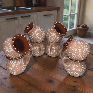 Pode incluir: Um conjunto de seis canecas de cerâmica bege com um padrão floral branco. Cada caneca tem uma borda e uma alça marrons. As canecas estão empilhadas em uma mesa de madeira, mostrando seu design e textura. Adequadas para servir bebidas.