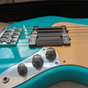 Puede incluir: Primer plano de un bajo eléctrico turquesa. El instrumento presenta un golpeador de madera clara, pastillas negras y herrajes cromados. El cuerpo tiene un acabado brillante y las cuerdas son visibles.