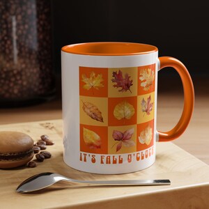 Könnte beinhalten: Weißer Keramikbecher mit orangefarbenem Innenraum und Griff. Der Becher zeigt ein Gitter aus bunten Herbstblättern auf orange-gelbem Hintergrund. Der Text "IT'S FALL O'CLOCK" ist auf der Vorderseite aufgedruckt.