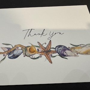 May include: A white card with the words "Thank you" in elegant script. The bottom of the card features a watercolor illustration of seashells, a starfish, and pearls, creating a coastal theme.