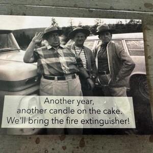 May include: Black and white photograph of three men in hats posing near a vintage car. The image includes the text: "Another year, another candle on the cake. We'll bring the fire extinguisher!"