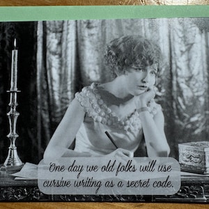 May include: A black and white greeting card featuring a vintage image of a woman writing. The card includes the text: "One day we old folks will use cursive writing as a secret code."