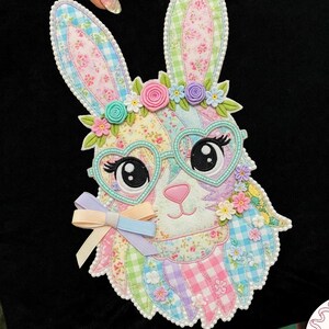 May include: Black pillow with a colorful patchwork bunny design. The bunny has heart-shaped glasses, floral accents, and a bow. The design incorporates pastel colors and floral patterns, creating a whimsical and decorative item.