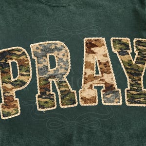 Könnte beinhalten: Dunkelgrünes Sweatshirt mit dem Wort "PRAY" in großen Buchstaben mit Tarnmuster. Die Buchstaben sind mit einem beigefarbenen, genähten Rand umrandet. Das Sweatshirt hat einen Rundhalsausschnitt und kurze Ärmel.