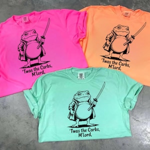 May include: Three t-shirts in neon pink, orange, and mint green, each featuring a black illustration of a frog in a coat holding a sword. The text "'Twas the Carbs, M'Lord," is below the frog.